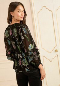 Černá blůza s propracovanými květinovými vzory v zelené, růžové a modré. Má volánové rukávy a peplum design. Nosit s tmavými džíny.