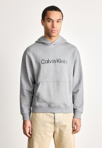Muž oblečený v světlém šedém mikině Calvin Klein a béžových kalhotách, stojící proti jednoduchému bílému pozadí a čelící dopředu.