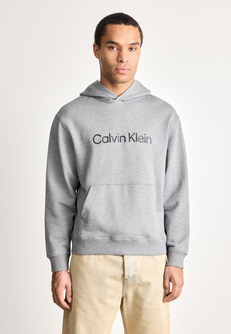 Muž oblečený v světlém šedém mikině Calvin Klein a béžových kalhotách, stojící proti jednoduchému bílému pozadí a čelící dopředu.