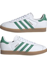 White sneakers with green accents, featuring three stripes and a textured gum sole. Branding "GAZELLE" on the sides. Classic athletic design.