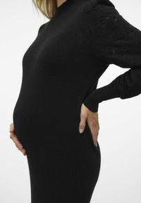 Schwarzes Strickkleid mit langen, strukturierten Ärmeln. Der Körper ist figurbetont und betont einen runden Bauch. Gerippter Halsausschnitt und Bündchen.