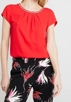 Mujer vistiendo una blusa de manga corta de color rojo brillante con un escote adornado con cuentas, combinada con pantalones negros con un estampado floral en rojo, blanco y rosa.