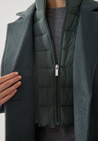 Cappotto di lana verde scuro con fodera imbottita trapuntata con zip. La texture è morbida e calda, con due tasche frontali e un colletto alto.