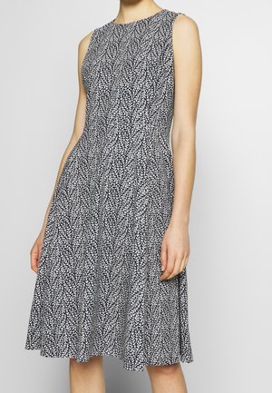 Ärmelloses schwarz-weißes knielanges Kleid mit taillierter Taille und blattähnlichem Muster, getragen von einer stehenden Person.