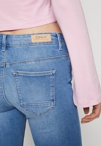 Vaqueros de denim azul claro con bolsillos traseros y una etiqueta de marca de cuero con la inscripción "ONLY." Llevados con una blusa de manga larga rosa.