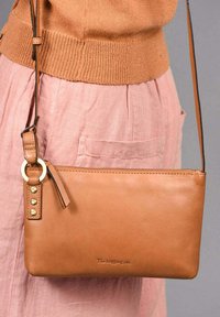 Sac crossbody en cuir tan avec une finition lisse, présentant un anneau de fixation en or et des clous sur la sangle. Fermeture éclair en haut.