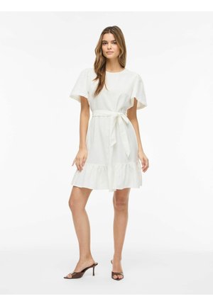 Žena stojaca, oblečená v bielych šatách po kolená s priedušnými rukávmi, pásikom v páse a hnedými otvorenými sandálmi na opätku.