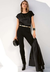 Schwarzes Satin-Oberteil mit kurzen Ärmeln, hochgeschnittene schwarze Skinny-Jeans, goldener Gürtel, schwarze High Heels, hält eine schwarze Tasche, schwarze Lederjacke hängt.