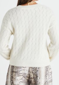 Maglione a maglia color crema con un motivo a zigzag testurizzato, con maniche lunghe e scollatura rotonda. Abbinato a pantaloni stampati.