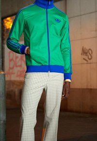 Vihreä vetoketjullinen takki sinisin yksityiskohdin ja raitoineen; ruudulliset housut beigeissä sävyissä. Sileä kangas, istuva malli ja rento urheilullinen tyyli.