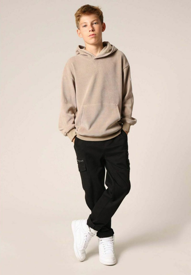 Hoodie beige texturé avec une poche kangourou, associé à un pantalon cargo noir avec des poches latérales et des baskets blanches.