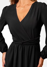 Vestido negro ajustado con mangas largas abullonadas, cinturilla acentuada, escote en V y textura de tela fluida. Collar dorado con colgante de estrella.