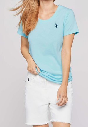 Mujer con camiseta de manga corta color azul claro y shorts vaqueros blancos con dobladillos deshilachados, de pie frente a un fondo liso.