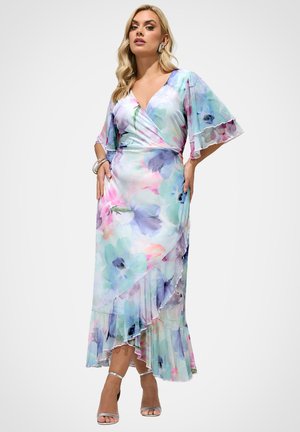 Kvinde i pastelblomstret omslagskjole med flagrende ærmer og flæsekant, iført sølvfarvede åbne hæle og sølvarmbånd mod hvid baggrund.