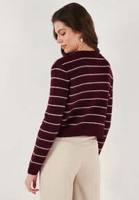 Burgundy kötött pulóver fehér vízszintes csíkokkal, cropped fazon, kerek nyakkivágás és hosszú ujjak, bézs nadrággal párosítva.