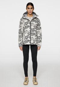Biała i czarna kurtka w wzory z kapturem, zamek błyskawiczny i długie rękawy, w połączeniu z czarnymi legginsami i beżowymi butami.