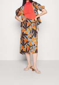 Jupe à motifs floraux colorés et haut assorti ; haut en tricot ribbed rouge, à manches courtes, avec un détail à nouer à la taille. Sandales à talons de couleur claire.