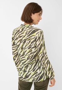 Lange mouw blouse van lichtgewicht stof met een beige basis en gedurfde groene en zwarte zigzagstrepen. Knopsluiting op de rug.