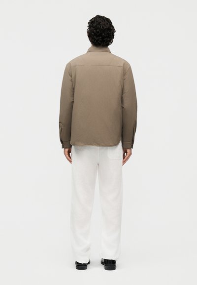 Άνδρας με σγουρά μαλλιά, που φοράει ανοιχτό καφέ σακάκι, άσπρο παντελόνι και μαύρα παπούτσια, στέκεται με την πλάτη προς το μέρος μας μπροστά σε ένα απλό φωτεινό φόντο.