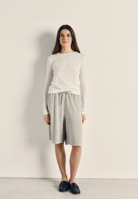 Žena stoji naslonjena na bež zid, nosi bijelu košulju dugih rukava, sive kratke hlače do koljena i crne mokasine.