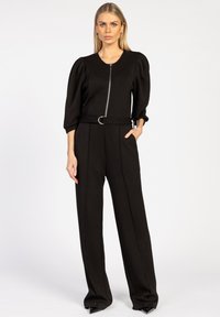 Zwarte jumpsuit met opgezette mouwen, een ritssluiting aan de voorkant en een ceintuur met een ronde gesp. Heeft rechte pijpen en een gladde stofstructuur.