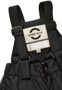 Schwarze wasserdichte Overalls mit verstellbaren schwarzen Trägern und einem cremefarbenen Etikett mit Markenlogo. Enthält Größeninformationen und Pflegehinweise.