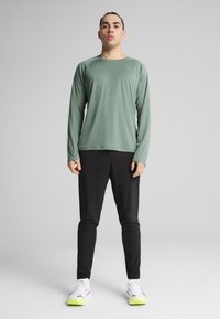 Hosszú ujjú, világoszöld sporting shirt, kerek nyakkivágással, fekete, szűk szabású nadrággal párosítva. Fehér sportcipők neon sárga részletekkel.