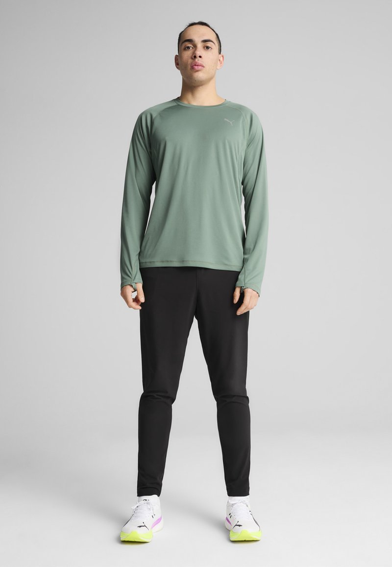 Hosszú ujjú, világoszöld sporting shirt, kerek nyakkivágással, fekete, szűk szabású nadrággal párosítva. Fehér sportcipők neon sárga részletekkel.