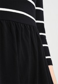 Schwarzes Kleid mit langen Ärmeln, versehen mit weißen horizontalen Streifen und einem gerafften Rockdesign. Hergestellt aus weichem, glattem Stoff.