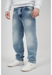Ljust blå denimjeans med en urtvättad finish, avslappnad passform och standard femfickorsdesign. Kombinerat med vita sneakers och en vit sweatshirt.