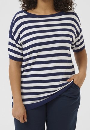 Person trägt einen marineblau-weiß gestreiften Kurzarm-Pullover und marineblaue Hose, eine Hand steckt in der Tasche, die andere entspannt neben dem Körper.