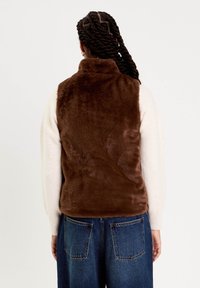 Personne avec des cheveux tressés portant un gilet en fausse fourrure marron sur un pull créme et un jean en denim bleu, vue de dos.