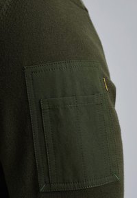 Dunkelgrüner Pullover mit einem strukturierten Strickkörper und einer flachen aufgenähten Tasche am linken Ärmel, mit kontrastierenden Nähten und einem Etikett.