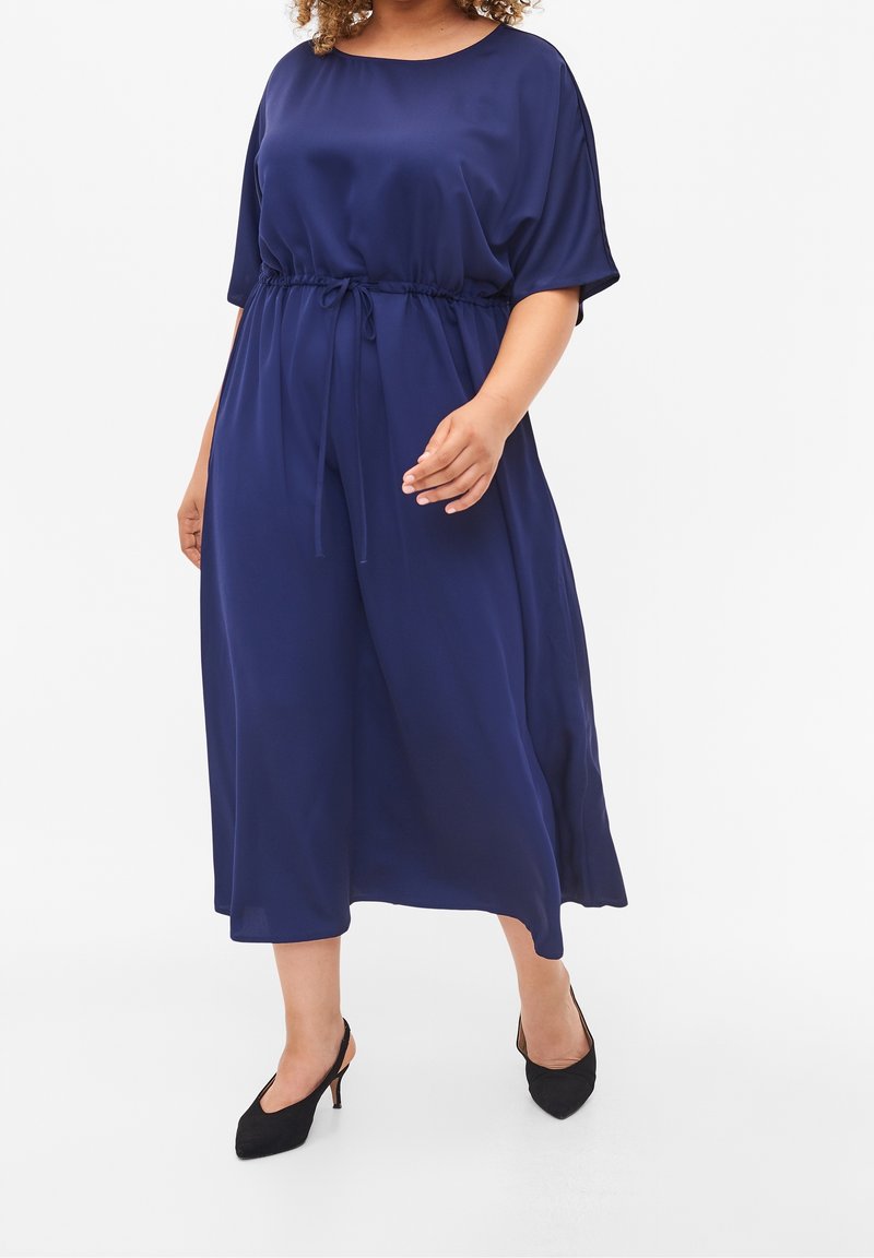 Femme portant une robe bleu marine ample, longueur genou, avec une ceinture nouée à la taille et des escarpins slingback noirs, sur un fond uni.