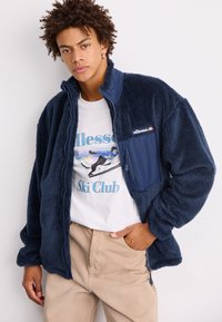 Tamno plava flis jakna sa patent zatvaračem, teksturiranim materijalom, visokim ovratnikom i kontrastnom glatkom najlonskom džepom. Nosena preko bijele majice s grafikom.