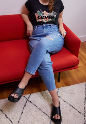 Sort grafisk t-shirt med blomsterdesign, lyseblå slidte jeans med tætsiddende pasform og sorte sandaler med krydset rem, siddende på en rød sofa.