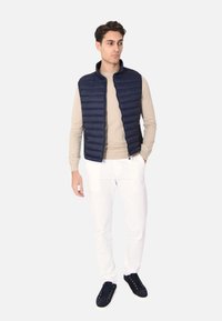 Gilet imbottito blu navy con colletto alto, tasche con zip e motivo trapuntato orizzontale, indossato sopra una camicia beige a maniche lunghe e pantaloni bianchi.