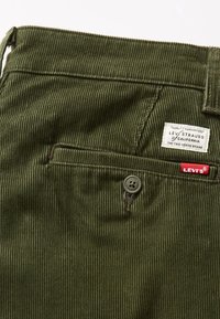 Primer plano de un bolsillo trasero de pantalones Levi's de pana verde con cierre de botón y dos etiquetas de marca encima del bolsillo.