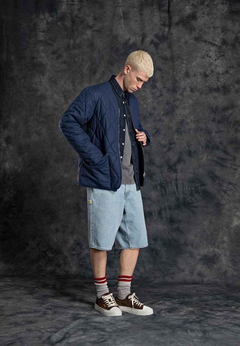 Chaqueta bomber acolchada de marinero sobre una camiseta gris, combinada con pantalones cortos de denim claros, calcetas grises con rayas rojas y zapatillas marrones con suelas blancas.