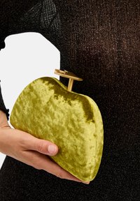 Sametti vihreä clutch, jossa on pyöristetty muoto, kullanvärisellä metallisella sulkijalla, pidetään kädessä tumman tekstuurin taustaa vasten.