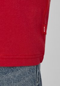 T-shirt en coton rouge à texture lisse, avec une étiquette latérale blanche indiquant "BZB". L'ourlet est soigneusement cousu. Porté avec un jean.