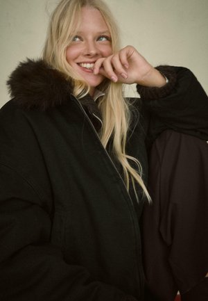 Crna oversize jakna s kapuljačom obloženom krznom. Ima prednji zip, velike džepove i udoban, teksturiran unutrašnji deo.