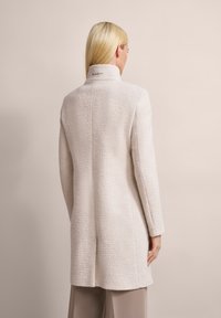 Helle beige, oversized Jacke aus strukturiertem Stoff, mit hohem Kragen und Logo-Detail auf dem Rücken. Lange Ärmel und gerader Schnitt unterstreichen das minimalistische Design.