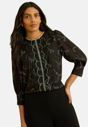 Frau mit langen dunklen Haaren, die eine schwarze transparente Bluse mit weißen Blumenmustern und gewelltem Saum trägt, kombiniert mit schwarzen Hosen.