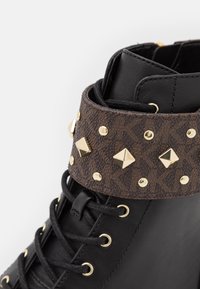 Czarny botek za kostkę z teksturowanym brązowym paskiem ozdobionym złotymi ćwiekami i piramidalnymi akcentami. Sznurowadła i przelotki w tym samym czarnym kolorze.