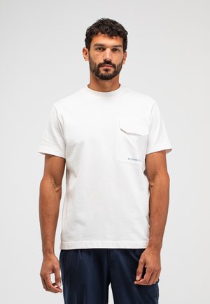 Man met kort krullend haar en baard, draagt een wit T-shirt met korte mouwen en een borstzak, en donkerblauwe broek, staand tegen een effen achtergrond.