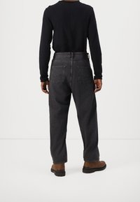 Persona con jeans negros holgados, camiseta de manga larga negra y botas marrones de pie sobre un suelo blanco contra un fondo blanco.