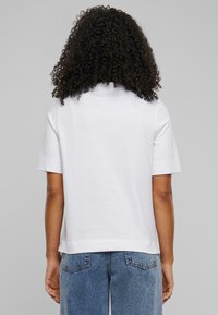 Hvit t-skjorte med korte ermer, rett passform og myk tekstur, kombinert med lyseblå denimjeans. Bakview som viser hårstruktur.