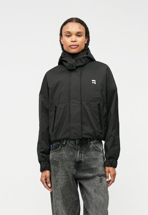 Black lightweight jacket with a hood, featuring a zip front, two zippered pockets, and a small logo on the chest. Textured fabric and elastic cuffs.