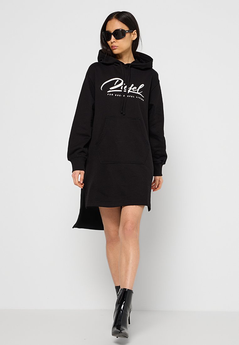 Vrouw in een zwarte hoodie jurk met een voorzak, zwarte zonnebril en zwarte enkellaarsjes, die tegen een effen achtergrond wandelt.